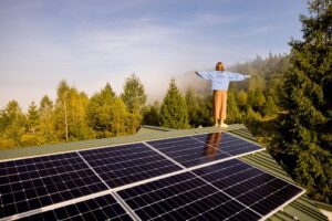 A woman stands on a solar-powered rooftop, arms outstretched, embracing the freedom and sustainability of off-grid living amidst a serene forested landscape