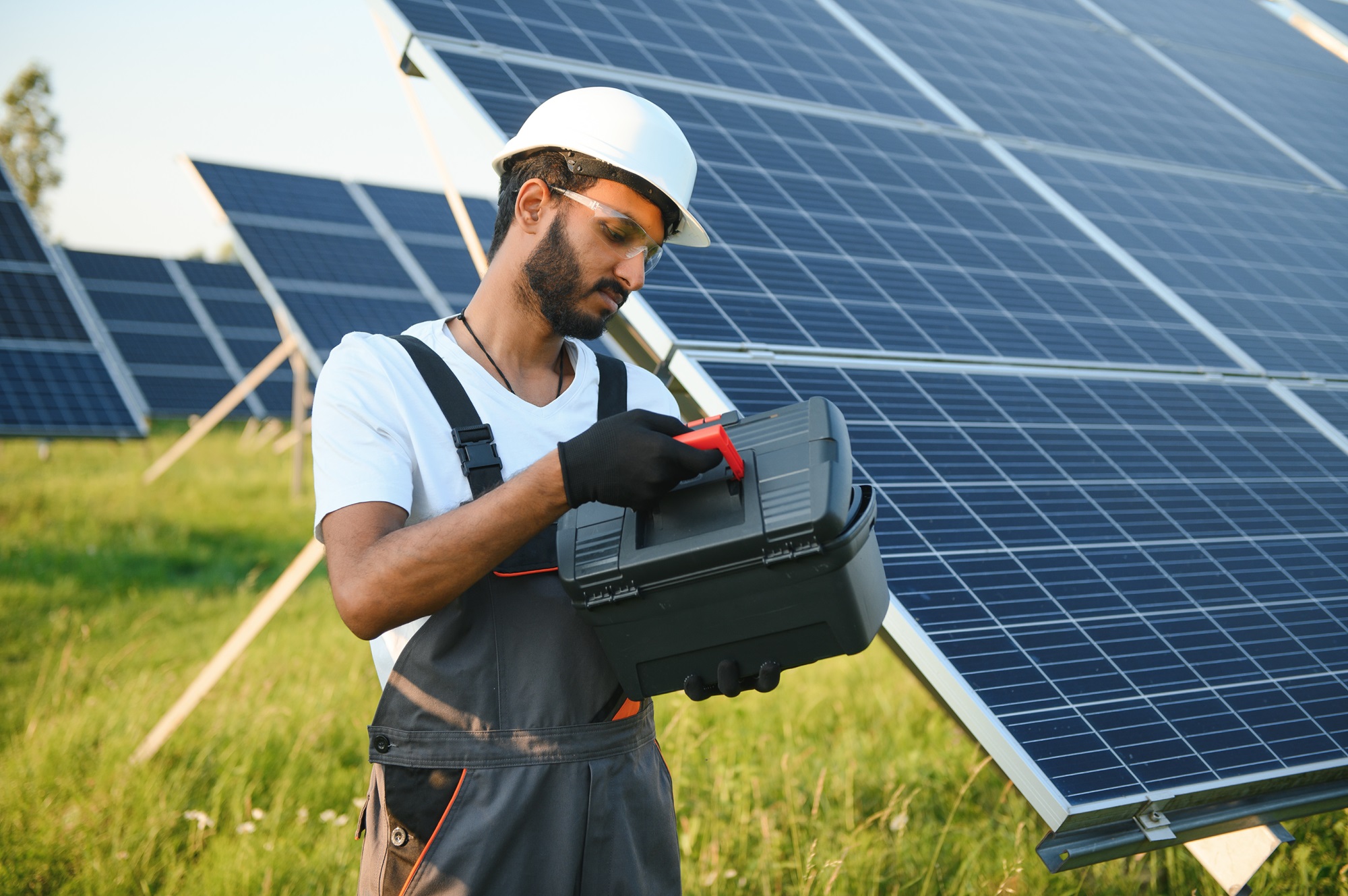 Male arabian engineer in helmet and brown overalls checking resistance in solar panels outdoors. Indian man working on station